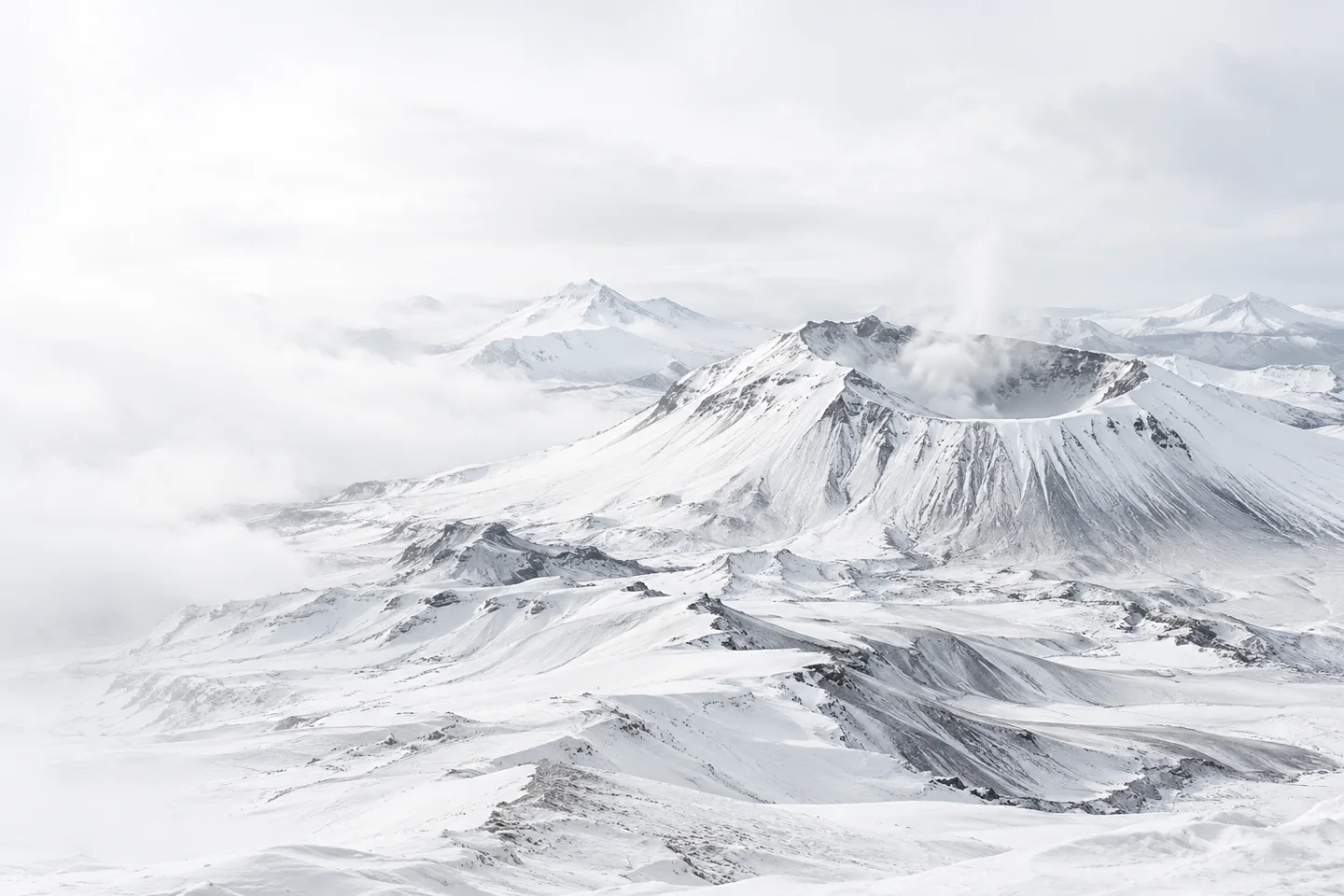A pale volcanic crater, surrounded by snow-covered peaks and finely textured ash ridges.